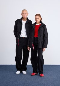 To personer står side om side iført sorte jakker, sorte bukser og sneakers i forskellige farver, mod en ensfarvet hvid baggrund.