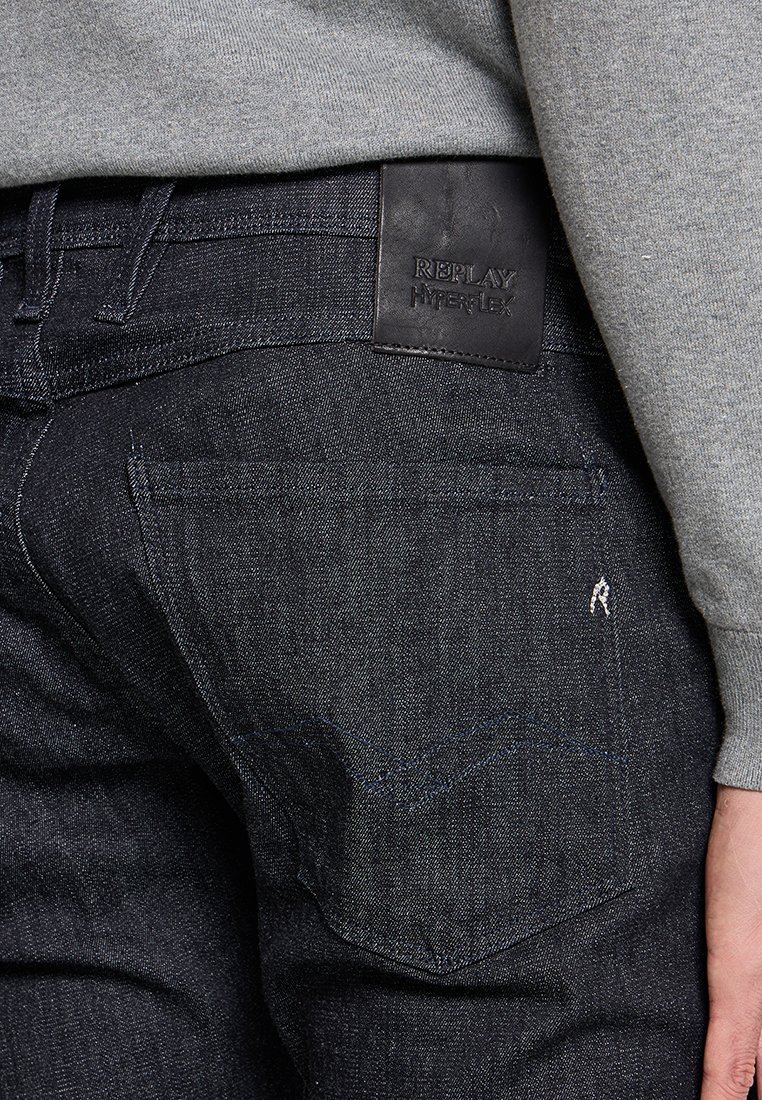 Dunkelblaue Denim-Jeans mit sichtbaren Gesäßtaschen, die mit einem Lederpatch mit der Aufschrift "REPLAY HYPERFLEX" versehen sind. Dezente Stickdetails im gesamten Design.
