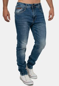 Blauwe denim jeans met een tapered fit, vervaagde was, vijf zakken en een merkpatch op de achterzak. Gecombineerd met witte sneakers.