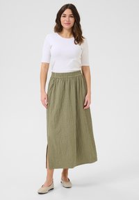 Olijfgroene, gestructureerde maxi rok met een elastische taille en een zijsplits, gecombineerd met een getailleerde witte top en lichte flats.