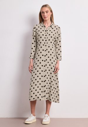 Femme portant une robe beige à manches longues boutonnée avec des pois noirs et des baskets blanches, debout contre un mur clair uni.