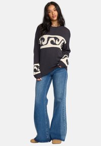Pullover di maglina navy con motivo a spirale crema, scollo rotondo e vestibilità oversize. Abbinato a jeans a gamba larga blu chiaro e calzature beige.