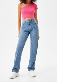 Kvinna i högmidjade blå jeans, vita sneakers och en rosa enfaldig kort topp som står mot en enkel vit bakgrund.