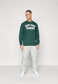 Grønn hettegenser med "NEW YORK YANKEES" trykket med hvitt, sammen med lysegrå cargo joggebukser og hvite sneakers.