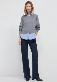 Maglione grigio a maglia sovrapposto a una camicia azzurra chiara, abbinato a pantaloni blu navy con dettagli in denim ai lati. Scarpe nere a punta completano l'outfit.