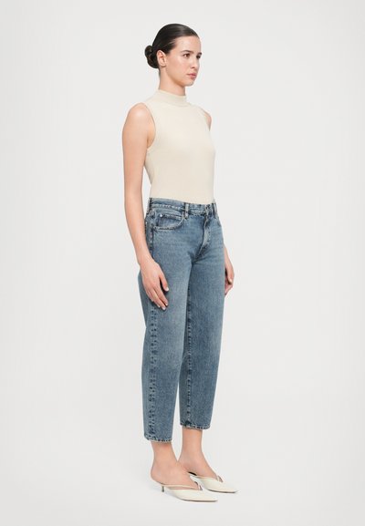 Beige mouwloze top, hoge hals; lichtblauwe high-waisted jeans met zijzakken; off-white puntige muiltjes; gladde textuur, ontspannen pasvorm.
