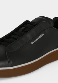 Μαύρο sneaker χαμηλού τύπου με καφέ σόλα, μαύρα κορδόνια και λογότυπο "Karl Lagerfeld" στο πλάι, εμφανίζοντας λεπτομερείς ραφές και υφή.