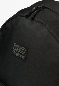 Zwarte rugzak met een gestructureerde stof, opvallende ritssluiting en een gestructureerd zwart label met de tekst "Superdry Outdoor Originals."