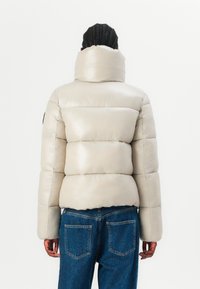 Pufferjack in lichtbeige met een glanzende afwerking, grote kraag en gestikt ontwerp. Gecombineerd met blauwe denim jeans.