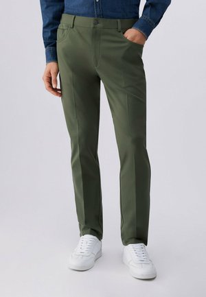 Muž v olivově zelených kalhotách rovného střihu, bílých teniskách a modré džínové košili stojící s jednou rukou v kapse.