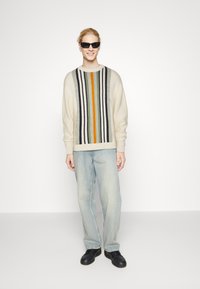 Pull crème avec des rayures verticales multicolores en vert, orange et marine, associé à un jean bleu clair et des chaussures noires.
