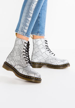 Vrouw draagt zilverkleurige veterlaarzen met slangenhuidpatroon, gele stiksels en doorzichtige zolen, gecombineerd met opgerolde blauwe jeans.