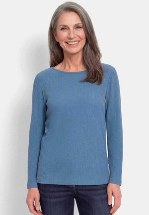 Femme aux cheveux gris souriante, portant un haut bleu côtelé à manches longues et un jean foncé, debout devant un fond clair uni.
