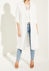 Manteau long blanc avec un large col et des manches retroussées, associé à un haut agrémenté de dentelle et un jean bleu clair. Sandales à talons hauts beige.