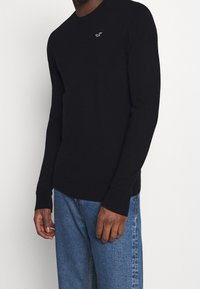 Svart stickad tröja med rundad hals och ribbade muddar. Innehåller en liten vit logotyp på bröstet. I kombination med ljusblå denimjeans.