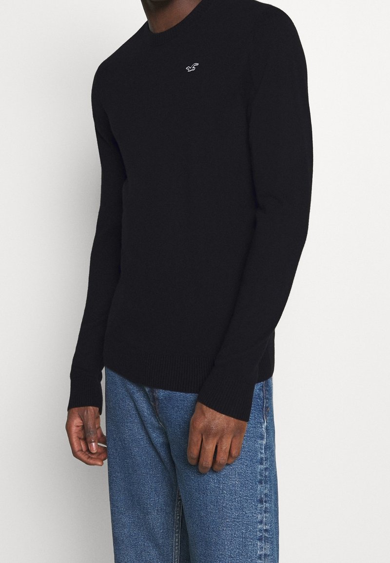 Svart stickad tröja med rundad hals och ribbade muddar. Innehåller en liten vit logotyp på bröstet. I kombination med ljusblå denimjeans.