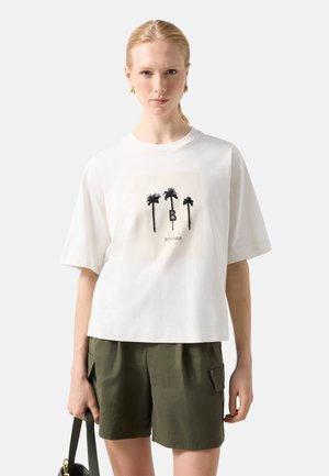 Kvinde iført hvid T-shirt med tre sorte palmer og teksten "Bogner", olivengrønne shorts, holder grøn håndtaske mod en lys baggrund.