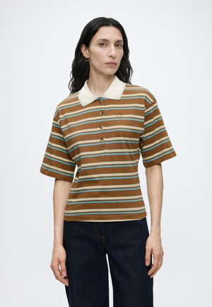 Mujer con cabello largo y oscuro viste una camiseta tipo polo de manga corta a rayas marrones y azules con cuello blanco y vaqueros azul oscuro, de pie sobre fondo blanco.
