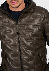 Man wearing a dark brown quilted leather jacket with a black hood and black zipper, shown from the chest up against a white background.