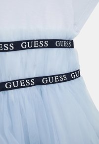 Jasnoniebieska sukienka z tiulu z dwoma czarnymi elastycznymi pasami w talii, na których powtarzają się białe logo "GUESS".