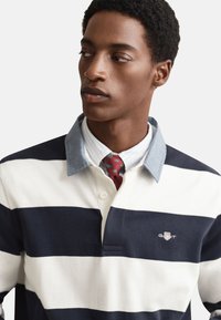 Poloshirt s mornarsko modro in belo črtasto vzorcem ter svetlim jeansa ovratnikom, z logotipom na levi strani prsnega koša, oblečen preko srajce s tankimi črtami in kravate.