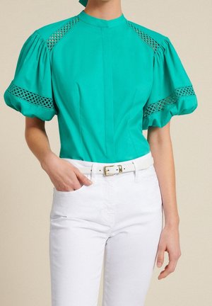 Blouse bleu sarcelle à manches bouffantes avec découpes ajourées en treillis, associée à un pantalon blanc taille haute et une ceinture blanche à boucle dorée.