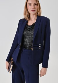 Marineblazer mit metallischen Akzenten, kombiniert mit einem schwarzen, strukturierten Oberteil. Slim-Fit-Hosen mit seitlichem Design. Clutch in der Hand.