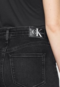 Los jeans de mezclilla negros presentan un diseño clásico de cinco bolsillos, con una etiqueta visible de Calvin Klein en la cinturilla y detalles de costura sutiles.