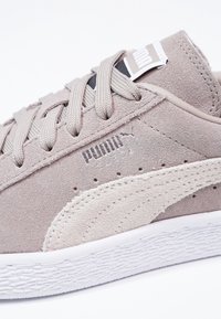 Beige Wildleder Sneaker mit strukturierter Oberfläche, versehen mit hellgrauen Akzenten, flachen Schnürsenkeln und einer weißen Gummisohle. PUMA-Logo auf der Seite eingeprägt.