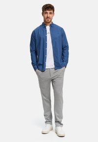 Man med händer i fickorna, iförd blå skjorta med knappar över en vit t-shirt, grå byxor och vita sneakers mot en enfärgad vit bakgrund.