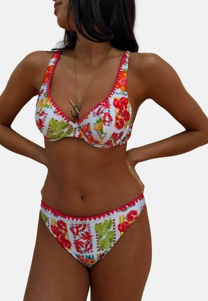 Donna che indossa un bikini bianco con motivi colorati di frutta e verdura, rifinitura rossa e una collana con ciondolo in oro.