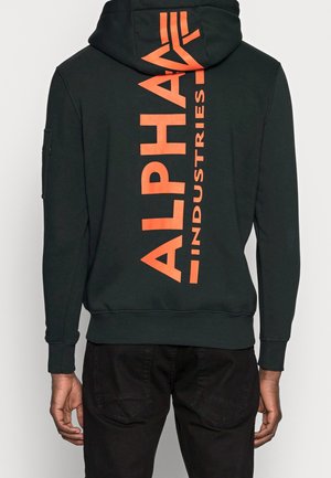 Czarna bluza z kapturem z pomarańczowym pionowym napisem "Alpha Industries" na plecach, noszona przez osobę stojącą tyłem do kamery.