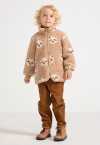 Fleece jacket in light brown with cat patterns, featuring a full zipper, high collar, and wide sleeves. Paired with brown corduroy pants and boots.