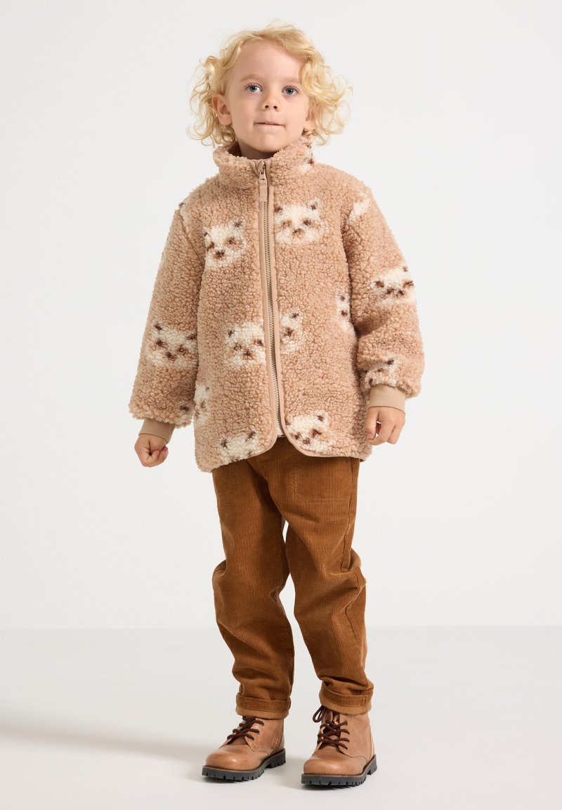 Fleece jacket in light brown with cat patterns, featuring a full zipper, high collar, and wide sleeves. Paired with brown corduroy pants and boots.