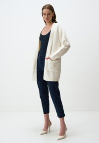 Cardigan bianco accogliente con apertura frontale, polsini a coste e tasche, abbinato a pantaloni blu navy e tacchi alti color crema. Tessuto liscio e vestibilità rilassata.