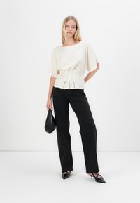Top color crema chiaro con maniche ampie e vita plissettata, abbinato a pantaloni neri a gamba dritta a vita alta e tacchi a punta neri lucidi.