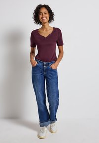 Bordový krátký rukáv top s výstřihem do V, spárovaný s modrými denimovými džíny. Džíny mají uvolněný střih a ohrnuté manžety. Bílé tenisky dokončují outfit.