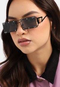 Mustat suorakulmaiset aurinkolasit, joissa on läpinäkyvät linssit, hopeiset niitit ja sawtooth-tyylinen yksityiskohta. Yhdistetty violettiin paitaan, jossa on musta kaulus.