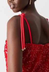 Robe en sequins rouges avec des bretelles arrondies et douces, nouées avec du tissu rouge et des accents en métal doré. Teint de peau lisse visible en dessous.