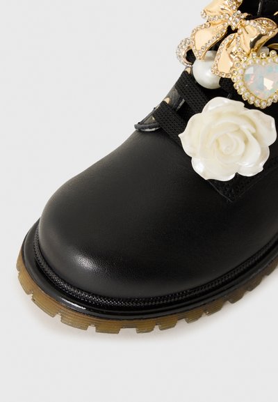 Bota de cuero negro con suela gruesa y translúcida, decorada con una rosa blanca y charm de mariposa dorada cerca de los cordones negros.