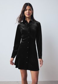 Černé sametové šaty s dlouhými rukávy, zapínáním na knoflíky vpředu, klasickým límečkem a rozšířeným střihem sukně. Hladká textura a jemný lesk.