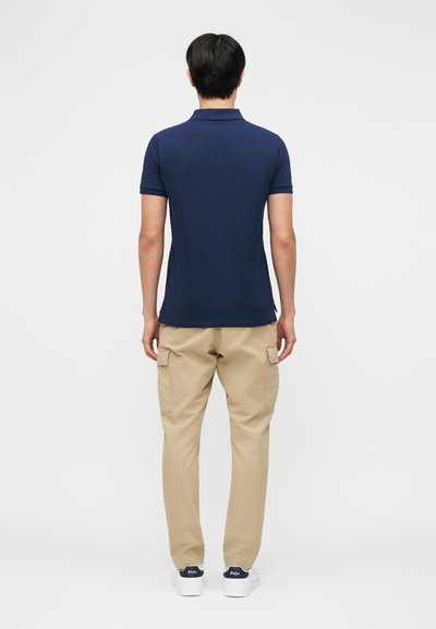 Uomo in piedi di spalle che indossa una polo blu navy, pantaloni cargo beige e scarpe da ginnastica bianche su uno sfondo chiaro uniforme.
