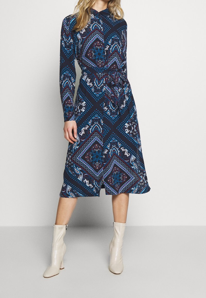 Femme portant une robe midi bleu marine à motifs avec des manches longues et des bottines blanches, debout devant un fond uni.