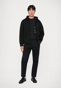 Svart zip-hoodie med en strukturert design og hette, brukt over en mørk skjorte, sammen med mørke bukser og svarte sko.