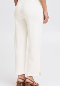 Pantalons blancs, légers, à jambes larges avec une taille élastique et des fentes latérales à l'ourlet, accompagnés de sandales à talons blocs à lanières brunes.