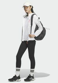 Chaqueta blanca ligera, leggings negros, zapatillas negras con detalles grises, gorra blanca, mochila negra y calcetines blancos con rayas.