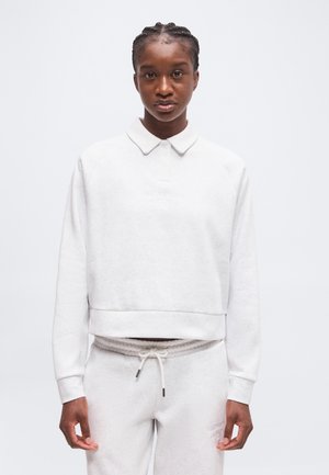 Femme aux cheveux tressés portant un sweat-shirt blanc à manches longues avec col et un pantalon de jogging blanc à cordon, debout devant un fond uni.