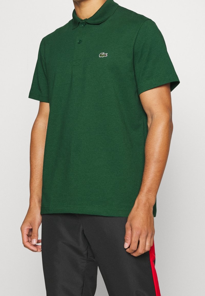 Homme portant un polo vert foncé à manches courtes avec un petit logo crocodile sur la poitrine et un pantalon noir avec une bande rouge.
