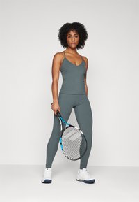 Sportswear set in muted green, featuring a fitted cami top with thin straps and high-waisted leggings. White athletic shoes and a tennis racket included.