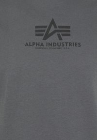 Pilkas medvilninis marškinėliai su juodu spausdintu "Alpha Industries" logotipu ir geometriniais akcentais. Lygi tekstūra, apvalus kaklo švarkelis.